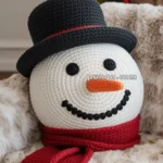 A crochet snowman pillow featuring a black hat, black button eyes, an orange carrot nose, and a red scarf, set against a cozy indoor background with blurred Christmas lights and decorations.