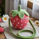 A crocheted strawberry-shaped crossbody bag with green leaves and a matching strap on a wooden table, surrounded by white daisies.