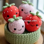 A knitted bowl containing four crocheted strawberries with smiling faces and green tops, placed on a wooden surface.