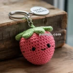 A crocheted pink strawberry keychain with green leaves and a smiling face, attached to a metal keyring with a round tag reading 'sweet treats,' resting on a wooden surface.