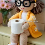 A crocheted doll wearing a yellow robe and slippers with daisy decorations sits on a cabinet, holding a roll of toilet paper. The doll has brown hair, black glasses, and is adorned with pink flowers in the background.