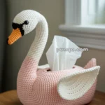 A knitted swan-shaped tissue box cover with a pink body and white neck, sitting on a wooden stool near a window.