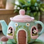 A crocheted teapot designed to resemble a whimsical house with a door, windows, and floral decorations, sitting on a crocheted base with a small mushroom next to it.