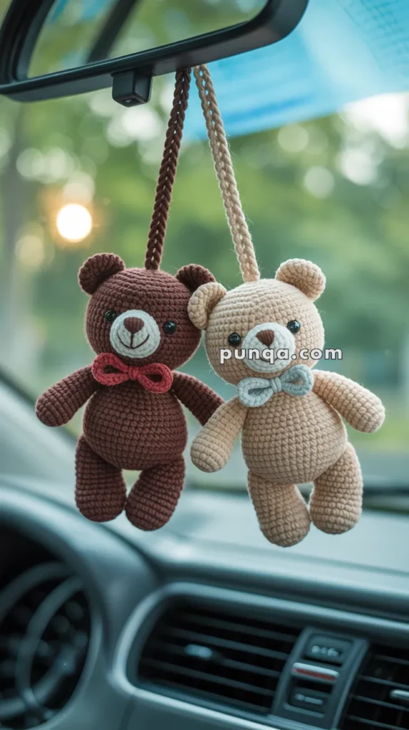 Two crocheted teddy bears, one brown and one beige, hanging from the rearview mirror of a car, each adorned with a small bow tie, against a blurred green background.