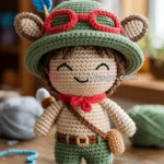 Crocheted doll with green hat and red goggles, featuring brown shorts, boots, and a blue feather, set on a wooden table.