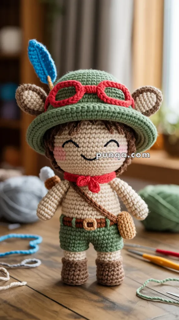 Crocheted doll with green hat and red goggles, featuring brown shorts, boots, and a blue feather, set on a wooden table.
