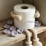 A roll of toilet paper with crocheted arms and legs sits on a wooden shelf, surrounded by small crocheted purple flowers.