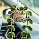 A crocheted hanging plant decoration with green leaves and a beige basket, suspended from the rearview mirror inside a car.