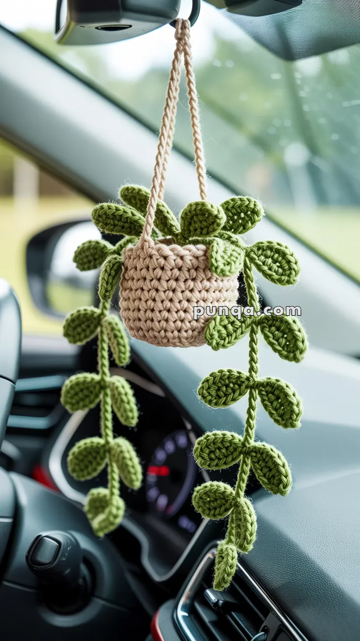 A crocheted hanging plant decoration with green leaves and a beige basket, suspended from the rearview mirror inside a car.