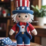 A crocheted doll dressed as Uncle Sam in a patriotic red, white, and blue outfit, featuring a top hat with stars, standing on a wooden table with an American flag and USA-themed decorations in the background.