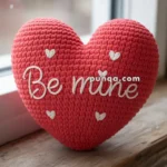 A knitted red heart-shaped pillow with the words "Be mine" stitched in white, displayed on a wooden windowsill.
