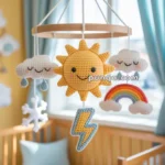A crocheted baby mobile featuring cheerful weather-themed elements including a smiling sun, clouds, raindrops, a rainbow, and a lightning bolt, hanging above a wooden crib in a sunlit nursery.