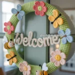 A crocheted wreath with colorful flowers, bees, and butterflies, featuring a "Welcome" sign, hung on a door.