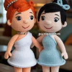 Two crocheted dolls standing on a wooden surface; one with orange hair styled in a bun and wearing a white dress, and the other with black hair in a bun and wearing a blue dress. Both are smiling and holding hands, with a home interior setting in the background.