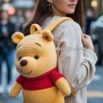 A young woman with brown hair is wearing a Winnie the Pooh backpack while standing on a city street. The backpack features a plush design with Pooh's iconic yellow body and red shirt. The background shows a blurred view of urban buildings and pedestrians.