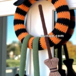 A crocheted wreath resembling a witch with a black and orange striped hat and legs, featuring black boots with yellow buckles and a brown broom hanging on a door.
