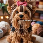 A handcrafted dog made from yarn, resembling a Yorkshire Terrier, with intricate details and a pink bow on its head, surrounded by various colored yarn balls on a woven mat.