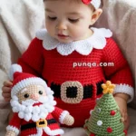 A baby dressed in a red crocheted Santa outfit with a white collar sits holding a crocheted Santa toy and a small crocheted Christmas tree, wearing a red bow headband.