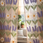 Crocheted curtains with lavender flowers and daisies, hanging in front of a sunlit room with potted plants and a white sofa in the background.