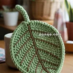 A crocheted green pot holder in the shape of a leaf with brown vein patterns, placed on a wooden surface amid potted plants in soft-focus in the background.