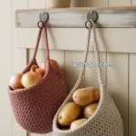 Two knitted hanging baskets filled with onions and potatoes, suspended on a rustic wooden shelf with a pot of lavender in the background.
