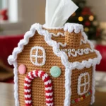A crochet gingerbread house tissue box cover with colorful decorations and a tissue emerging from the chimney, displayed on a wooden table with a blurred Christmas tree in the background.