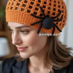 A person wearing an orange crocheted beanie adorned with a black knitted spider decoration, gazing to the side with a slight smile.