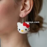 Close-up of a person wearing a crocheted earring shaped like a white cat with a red bow, with a website watermark reading "punqa.com."