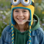 A smiling child wearing a crocheted hat designed to resemble a Minion, with yellow, blue, and black details, including large goggle-like eyes and ear flaps with braided tassels. The child is also wearing a green hoodie and a denim jacket, standing outdoors with a blurred natural background.