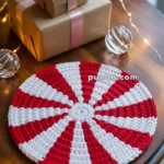 A red and white crocheted round mat resembling a peppermint candy on a wooden table, with wrapped gifts tied with pink ribbons and decorative string lights in the background.