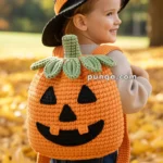 A young child wearing a witch's hat and a knitted pumpkin-shaped backpack stands amidst an autumn setting with leaves on the ground.