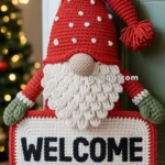 A crocheted gnome decoration with a red hat and green gloves holding a sign that reads "WELCOME" hangs on a door handle, with a blurred Christmas tree in the background.