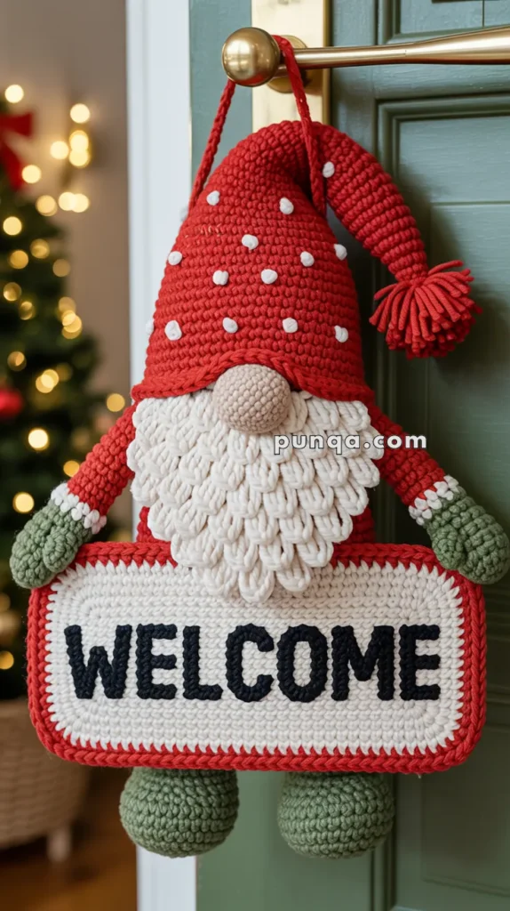 A crocheted gnome decoration with a red hat and green gloves holding a sign that reads "WELCOME" hangs on a door handle, with a blurred Christmas tree in the background.
