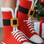 A pair of crochet boots designed to look like Santa's boots, featuring red, white, and black colors with a white sole and yellow buckle detail, worn by a person standing near a Christmas tree and gift.
