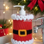 A soap dispenser adorned with a crocheted cover designed to resemble Santa's suit, featuring red, white, and black yarn with a decorative yellow buckle, placed on a bathroom sink with a festive wreath and fairy lights in the background.