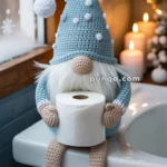 A crochet gnome figure wearing a blue hat with white pom-poms, holding a roll of toilet paper, sitting on the edge of a bathtub with candles and a snowy window in the background.