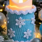 A candle wrapped in a blue crocheted sleeve adorned with white crocheted snowflakes, surrounded by Christmas decorations including pine branches, small lights, and ornaments.