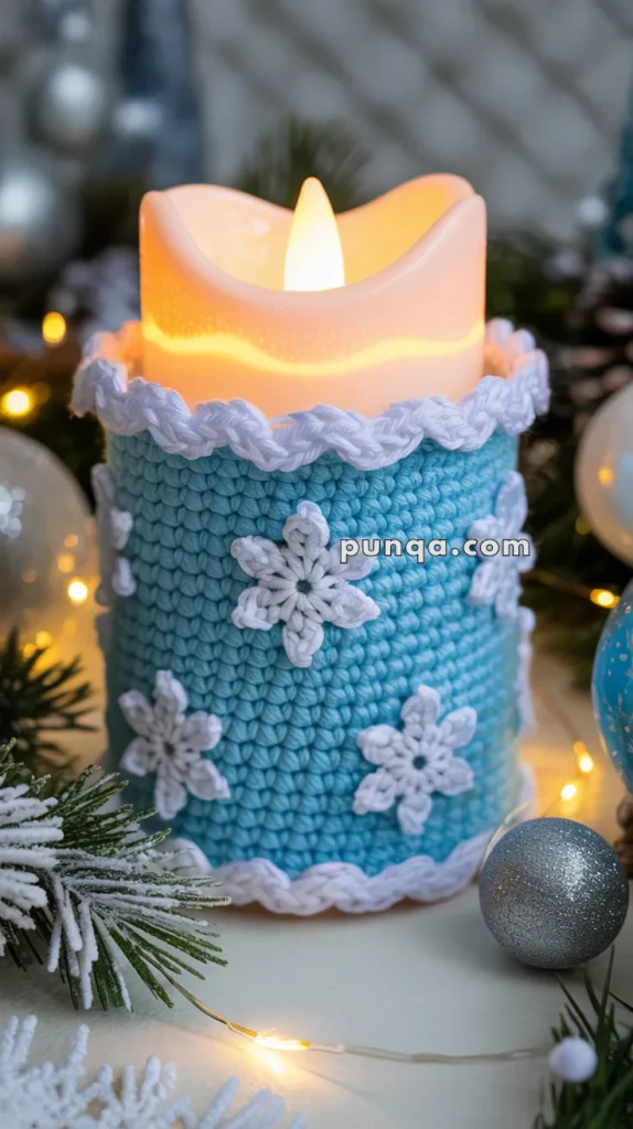 A candle wrapped in a blue crocheted sleeve adorned with white crocheted snowflakes, surrounded by Christmas decorations including pine branches, small lights, and ornaments.