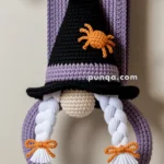 Crochet gnome decoration with a black witch hat, orange spider detail, and white braided hair, hanging on a towel rack above a white towel.